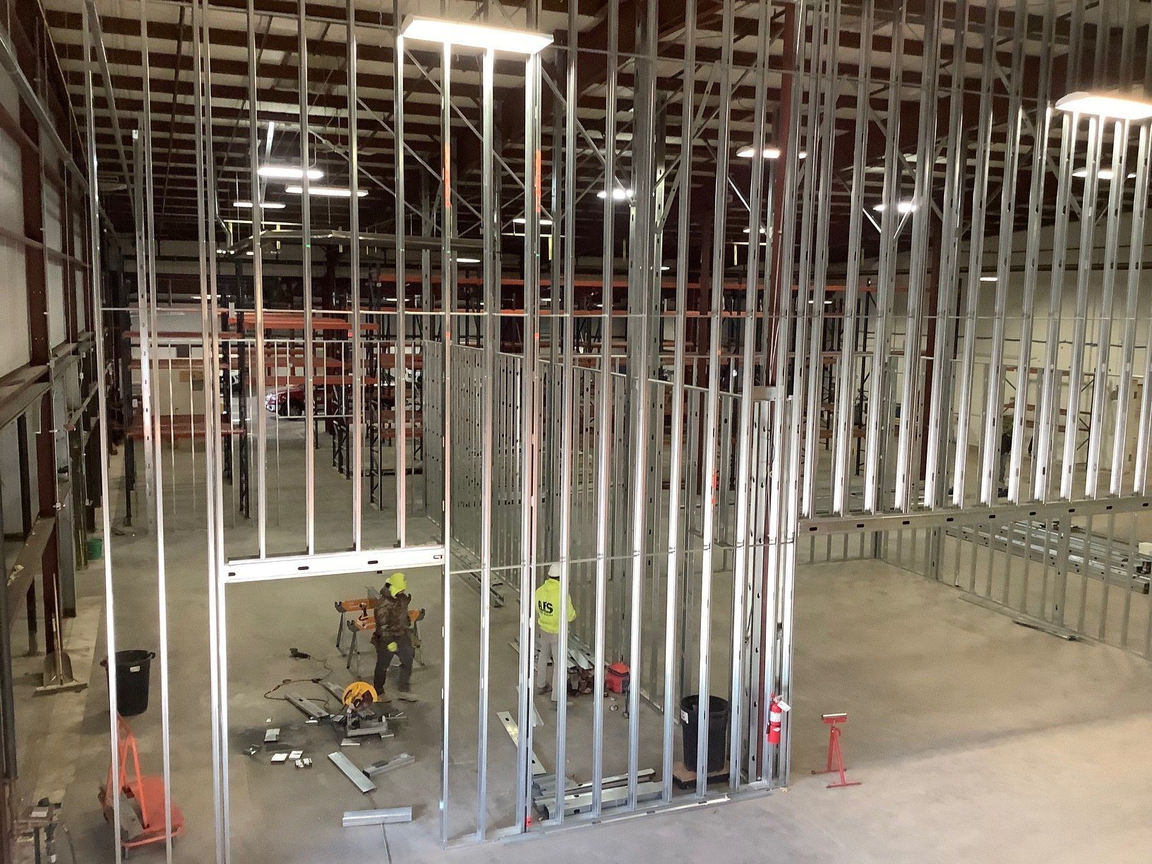 Interior view of a building under construction, metal framing for walls and workers in the distance.