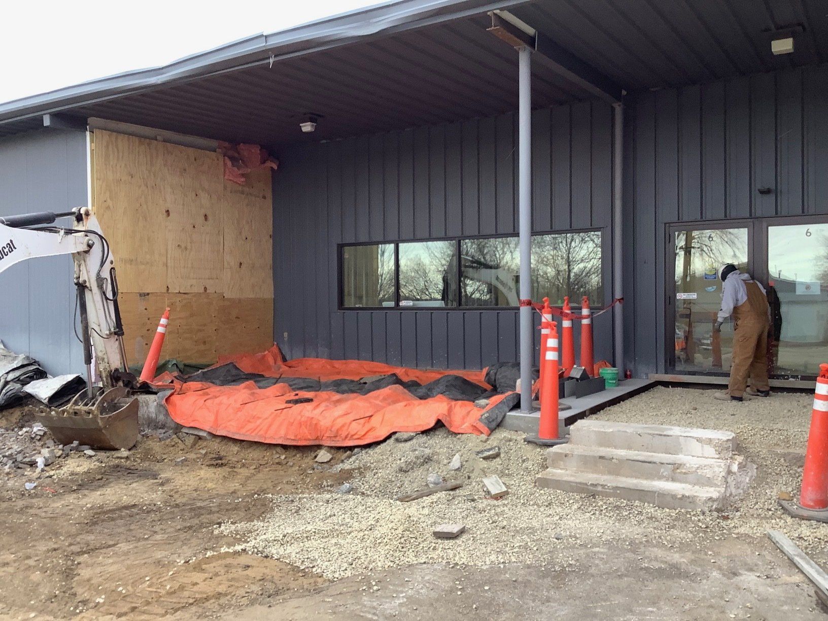 Construction site. Building with gray siding and plywood where wall removed. Excavator, debris, and worker present.