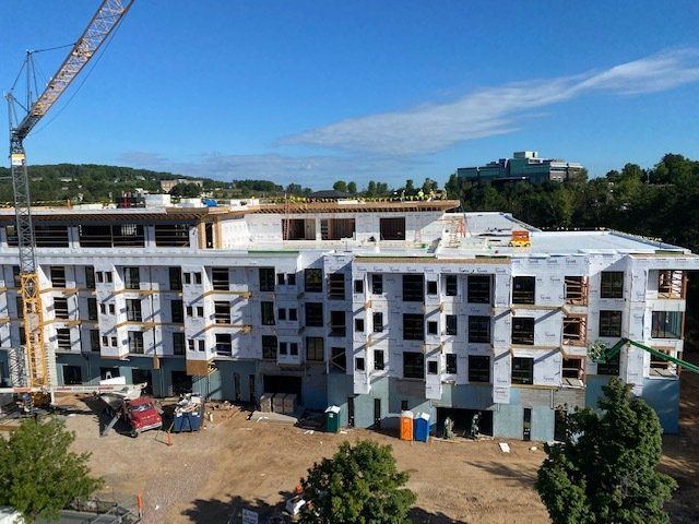 Construction site of a multi-story building, with a crane, surrounded by equipment, trees, and blue sky.