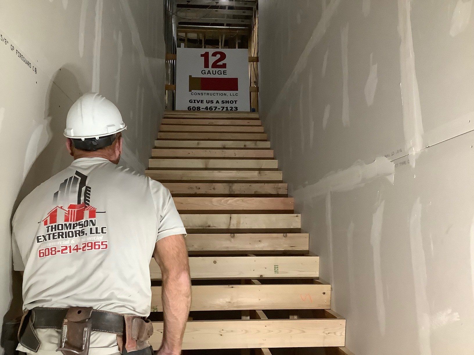 Construction worker looking up a wooden staircase in a building under construction. White hard hat, drywall walls.
