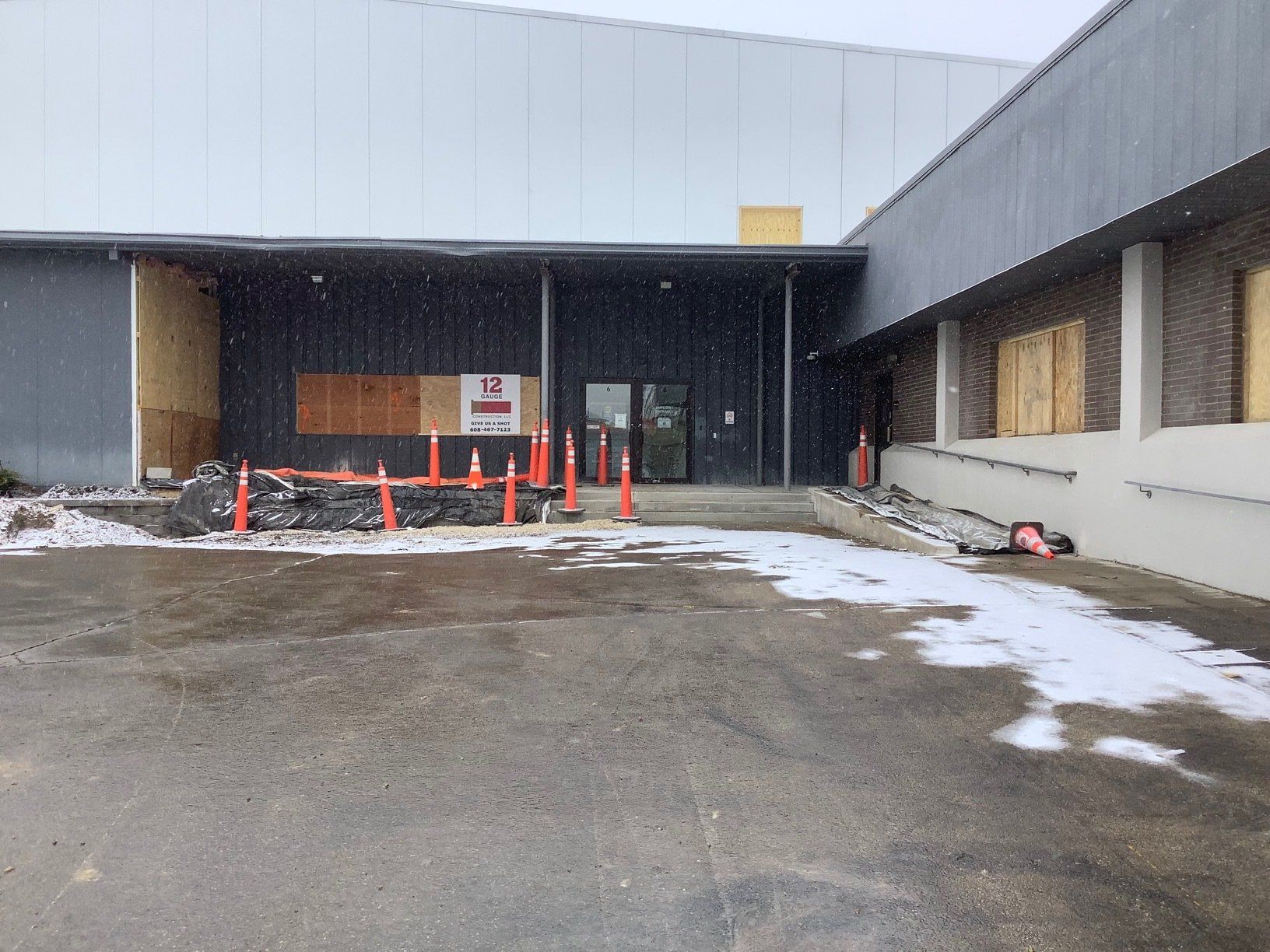 Exterior of a building with boarded-up windows and an entrance blocked by construction materials. Snow falling.