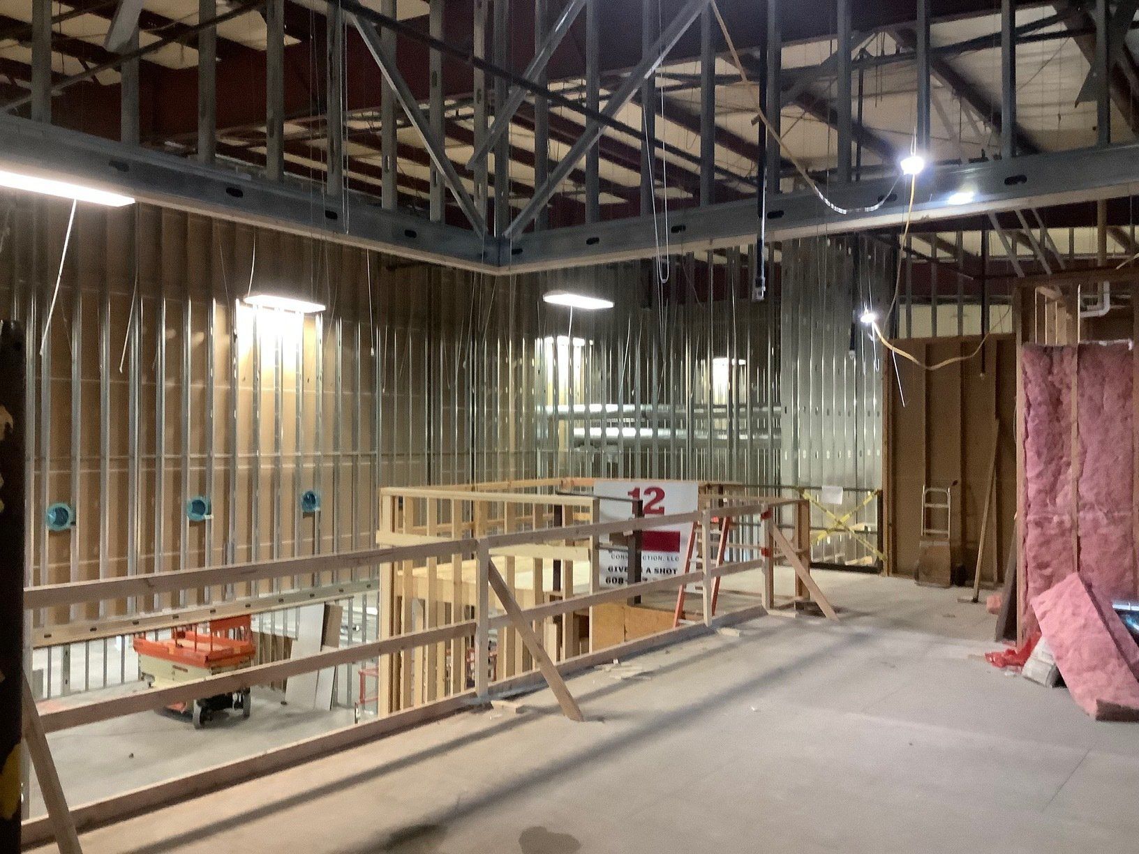 Interior view of a building under construction. Framing, exposed studs, and a partial second story with a railing are visible.