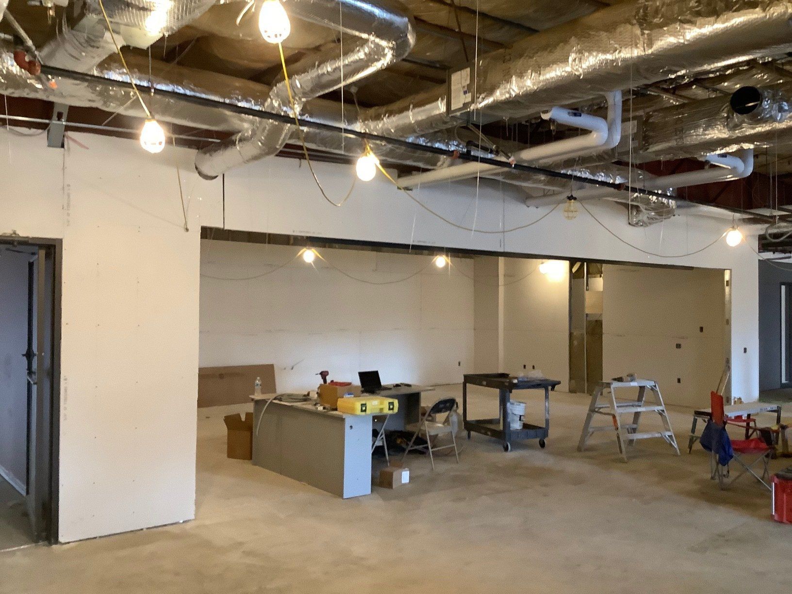 Interior of a room under construction with exposed pipes and wires. White walls, concrete floor, work tools visible.