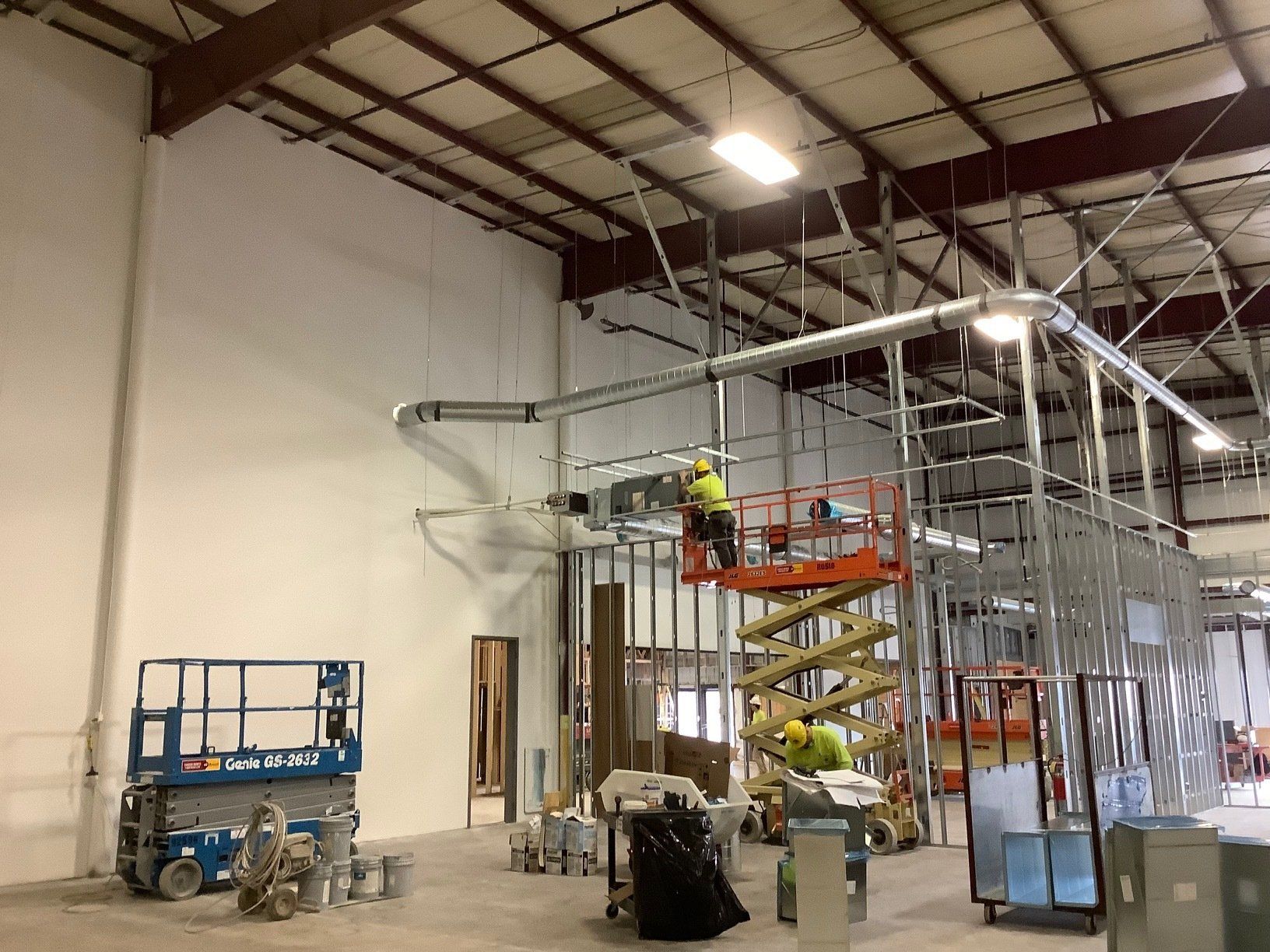 Construction site interior; workers on lifts installing ductwork and framing a wall.