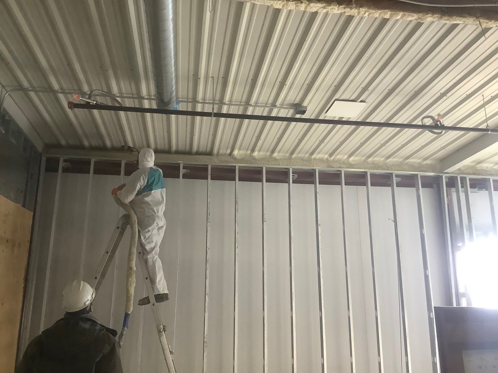 Two workers in protective suits spray insulation on a wall in a warehouse-like setting.