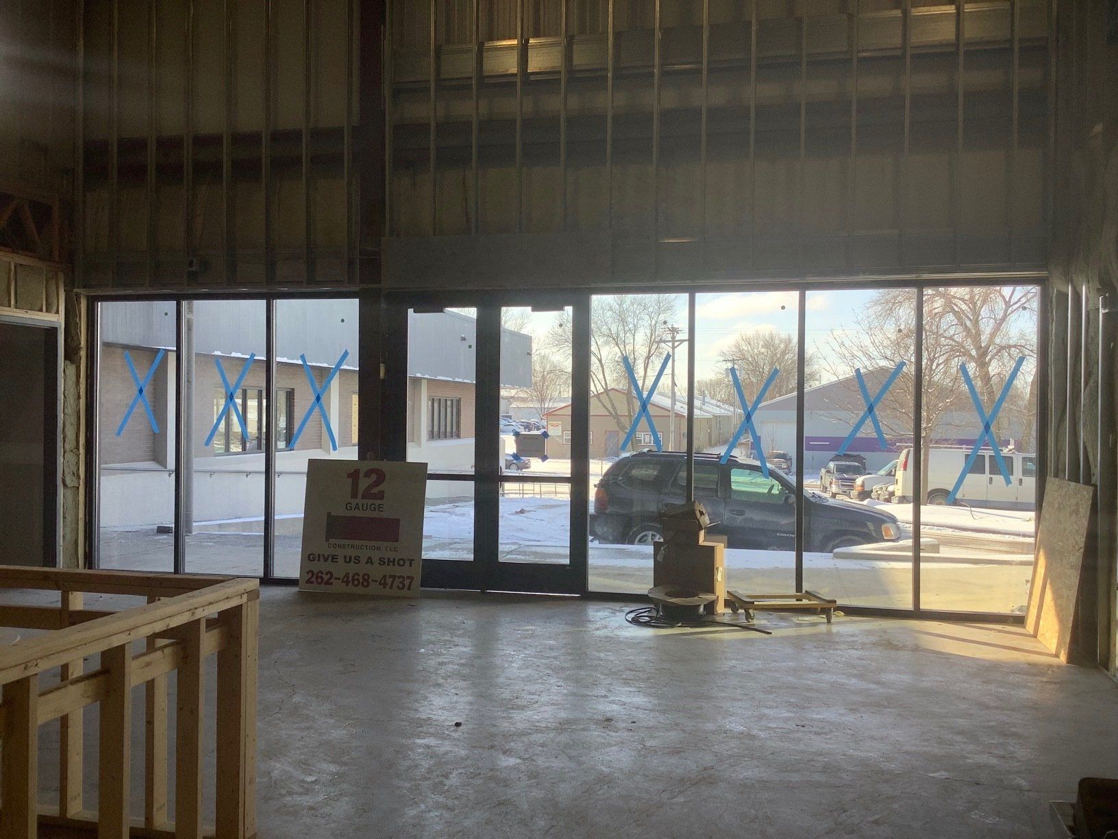 Interior of a building with large windows looking out onto a street with parked vehicles. Construction in progress.