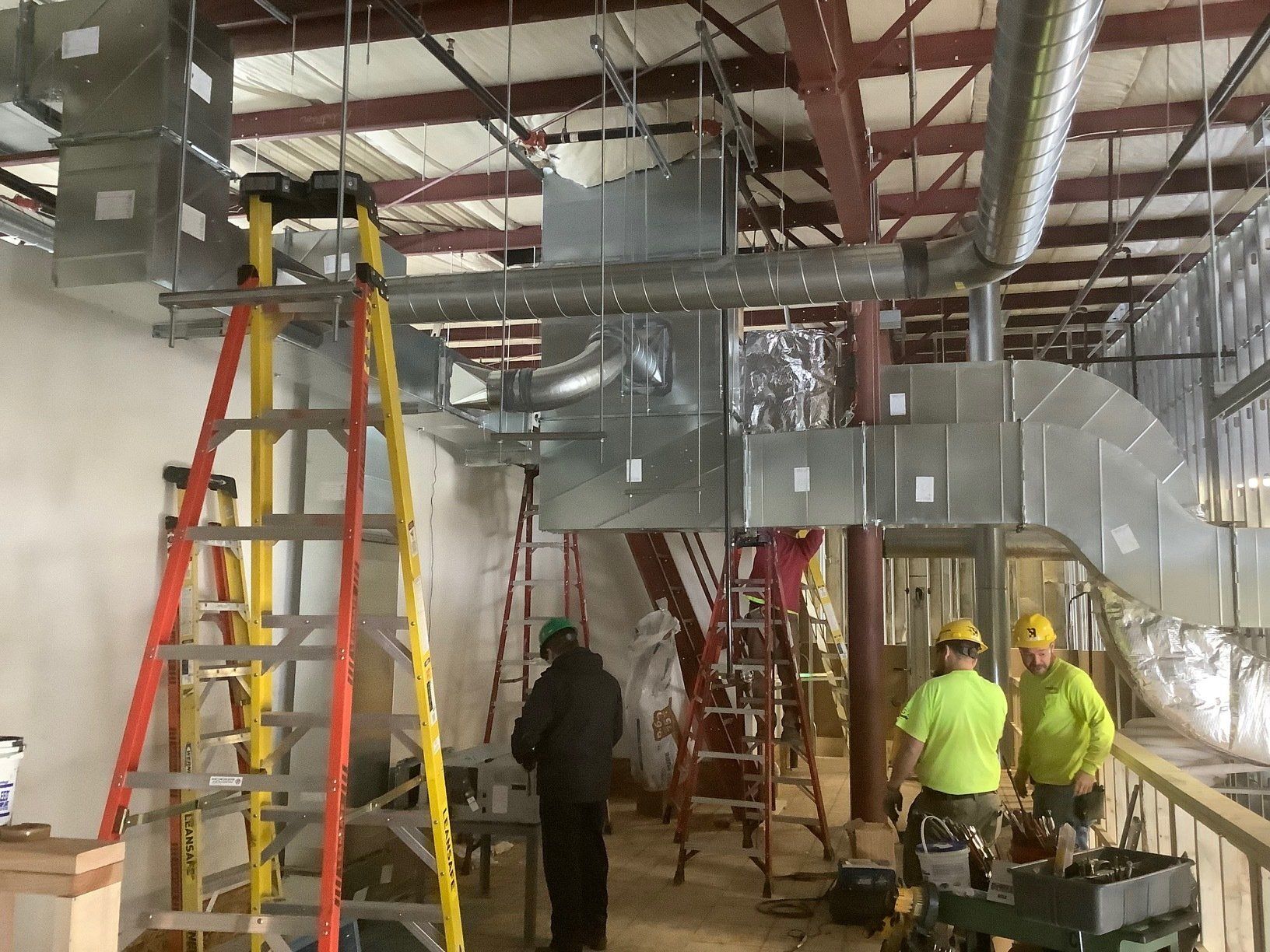 HVAC ductwork installation in progress; workers on ladders. Metallic ducts, exposed beams, and a partial building interior.