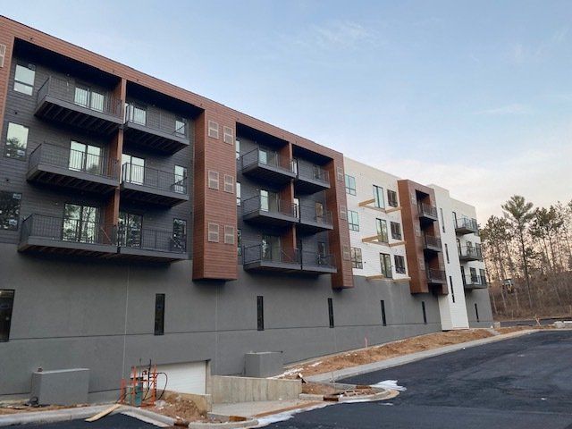 Modern apartment building exterior with balconies, gray, brown and white siding, and a sloped driveway.