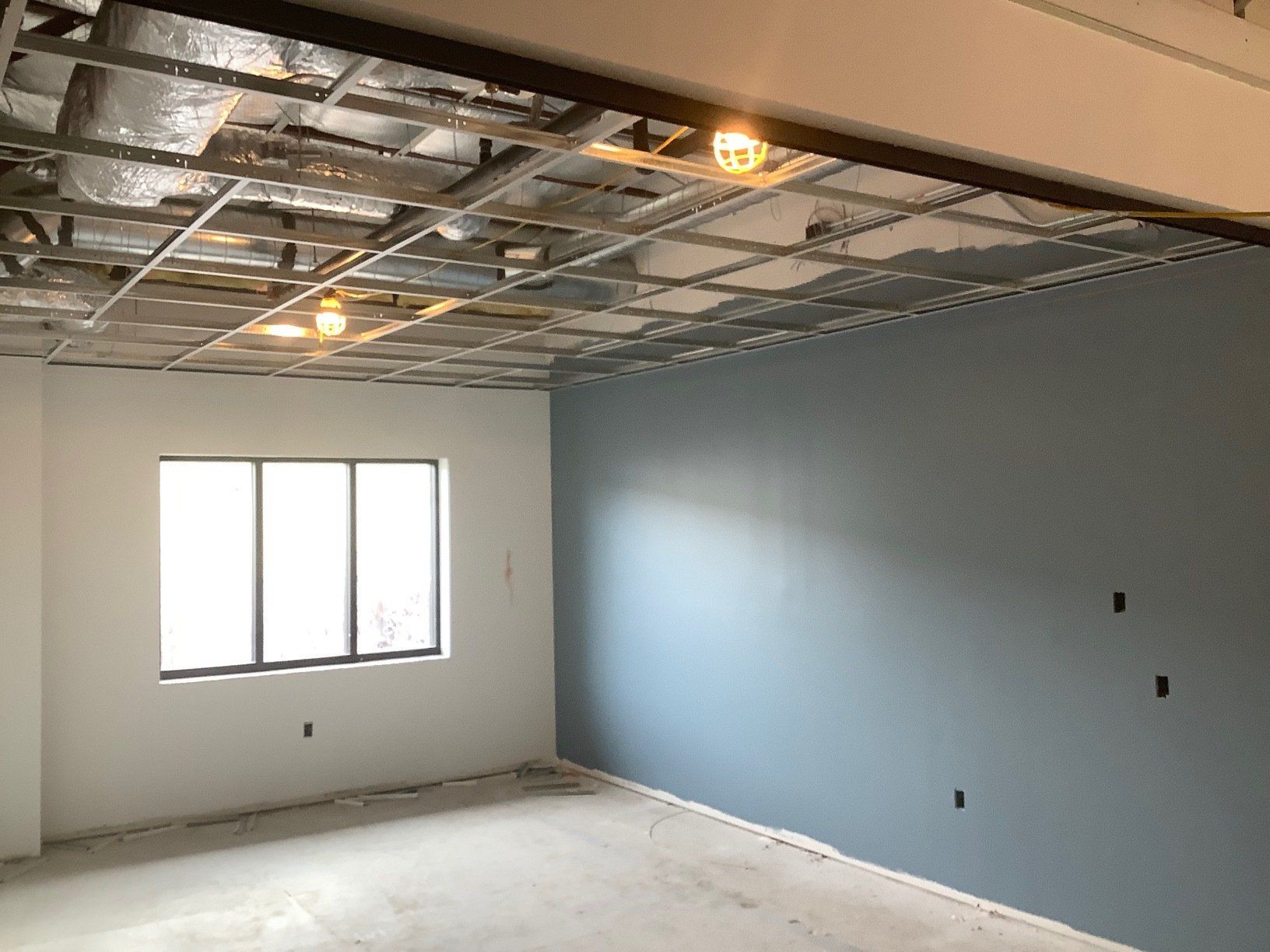 Room interior under construction with exposed ceiling grid, two-tone walls, and window.