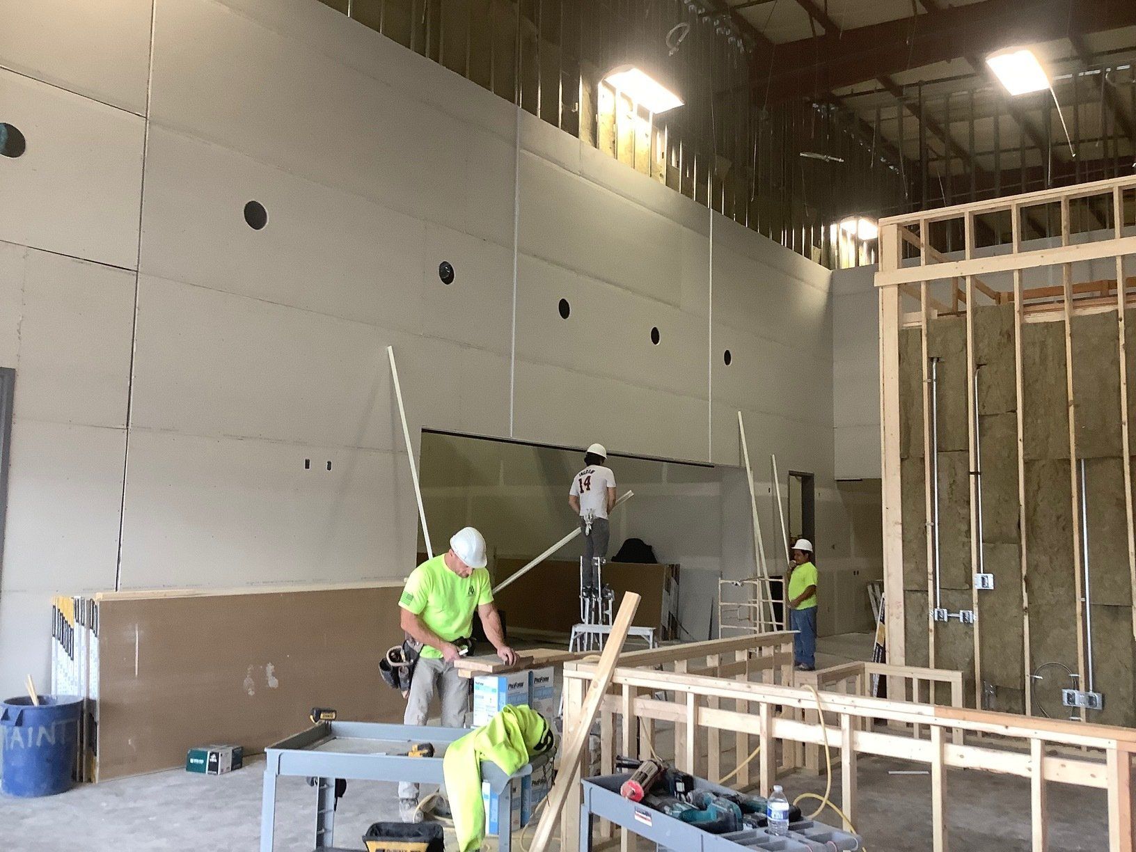 Construction workers installing drywall on a large interior wall, with exposed framing visible.