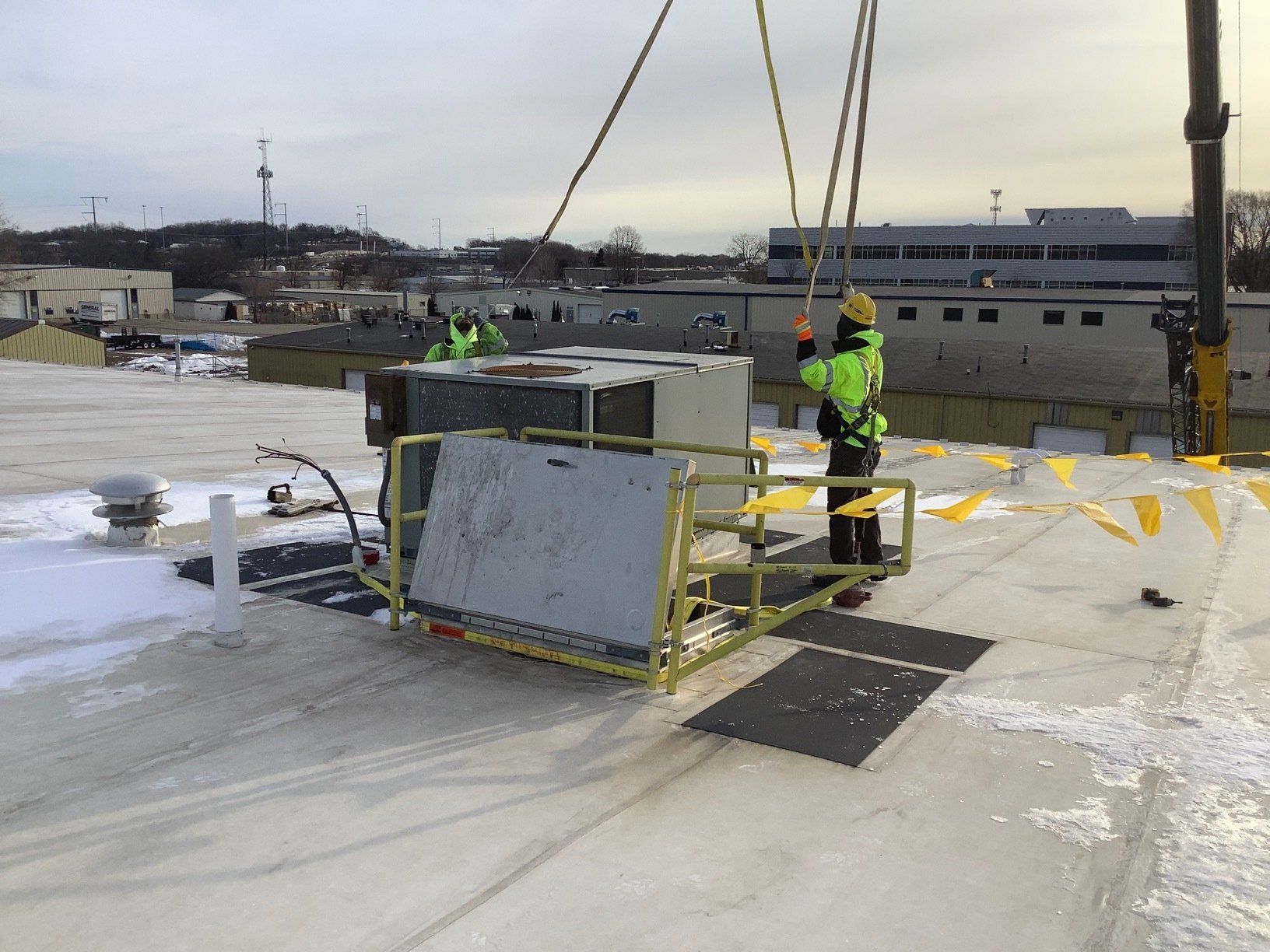 Workers installing HVAC unit on a rooftop using a crane. Two wear safety vests. Snowy conditions.