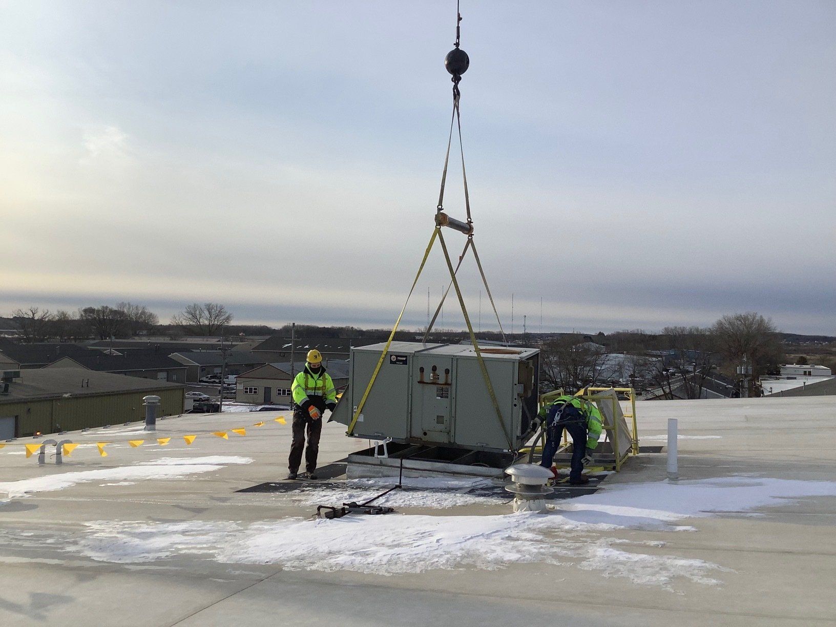 Two workers on a rooftop installing a large HVAC unit with a crane; snowy conditions.