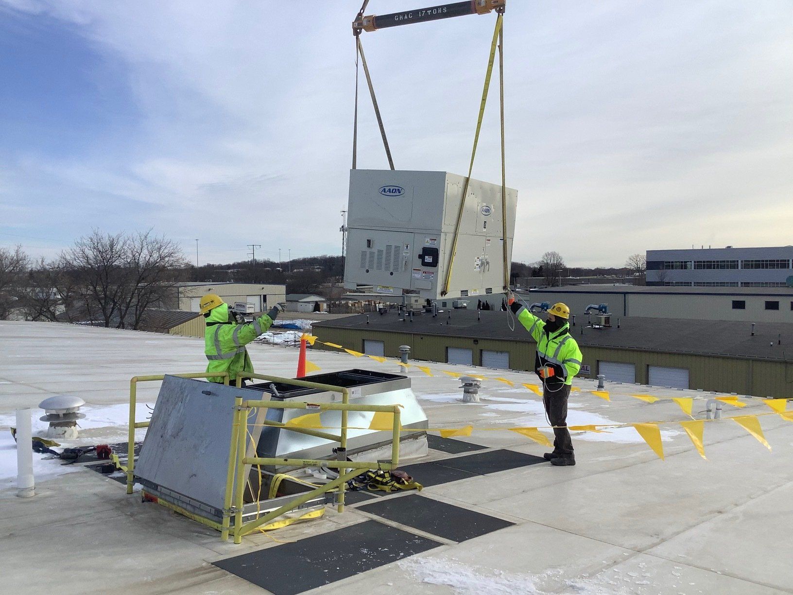 Two workers guiding a large, light-colored industrial unit being lifted by a crane onto a building rooftop.