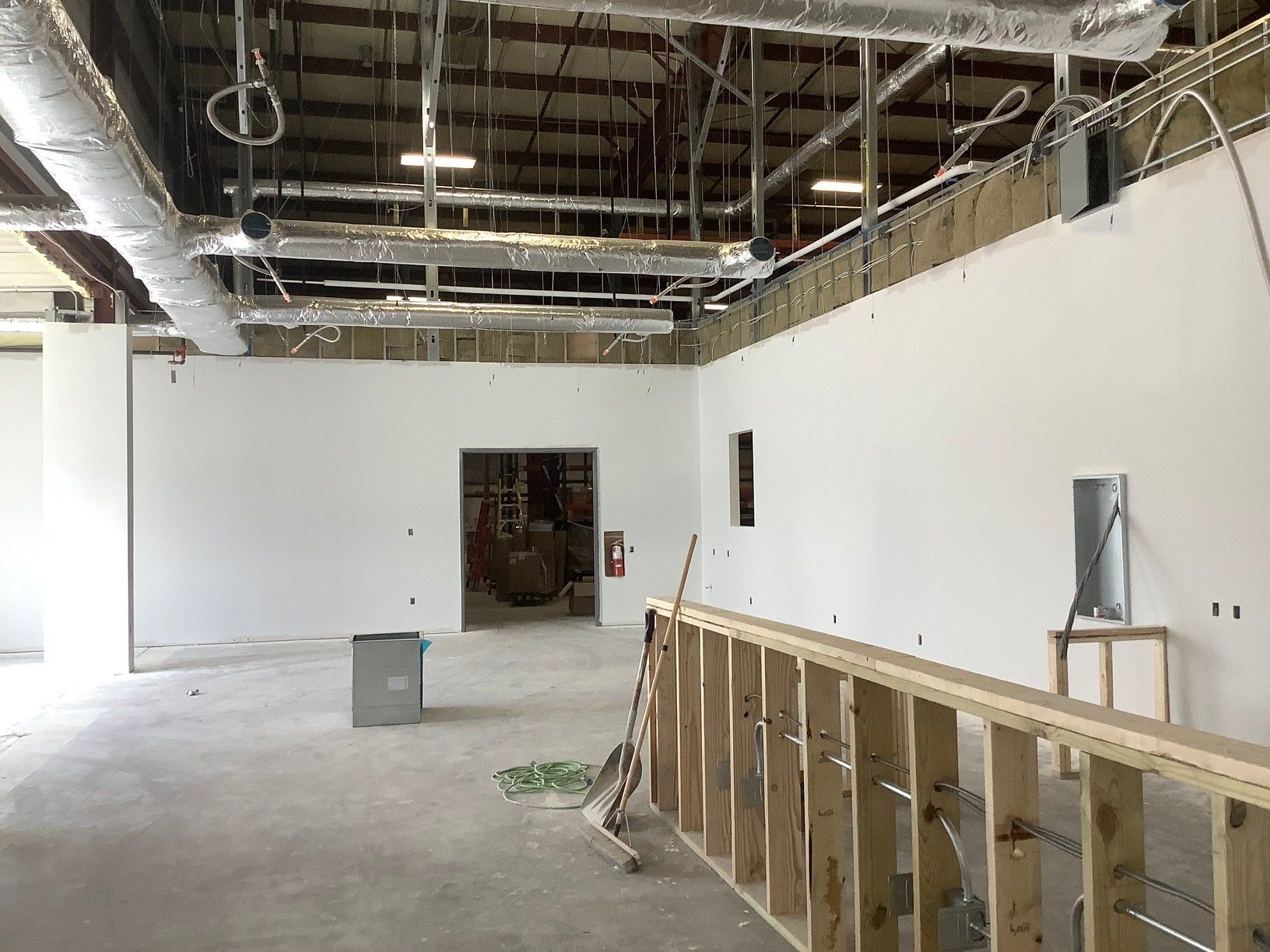 Interior of a building under construction. White walls, exposed ceiling supports, and a partially built wooden frame are visible.