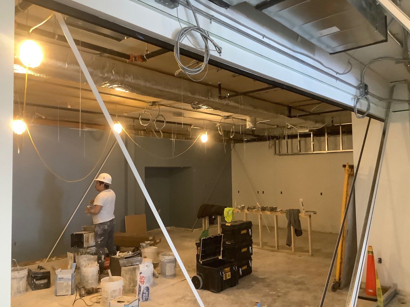 Interior of a construction site: ceiling pipes, exposed lights, a worker in a hard hat, and tools.