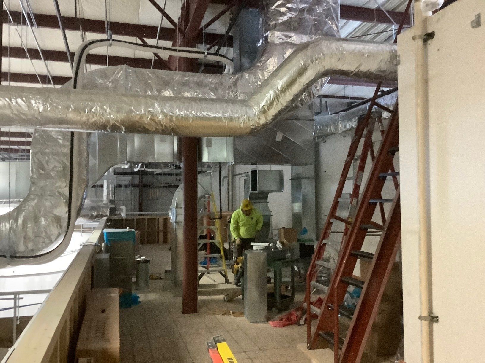 HVAC ductwork installation in an industrial space. Worker on the floor by equipment, red ladder, overhead pipes, and metal beams.