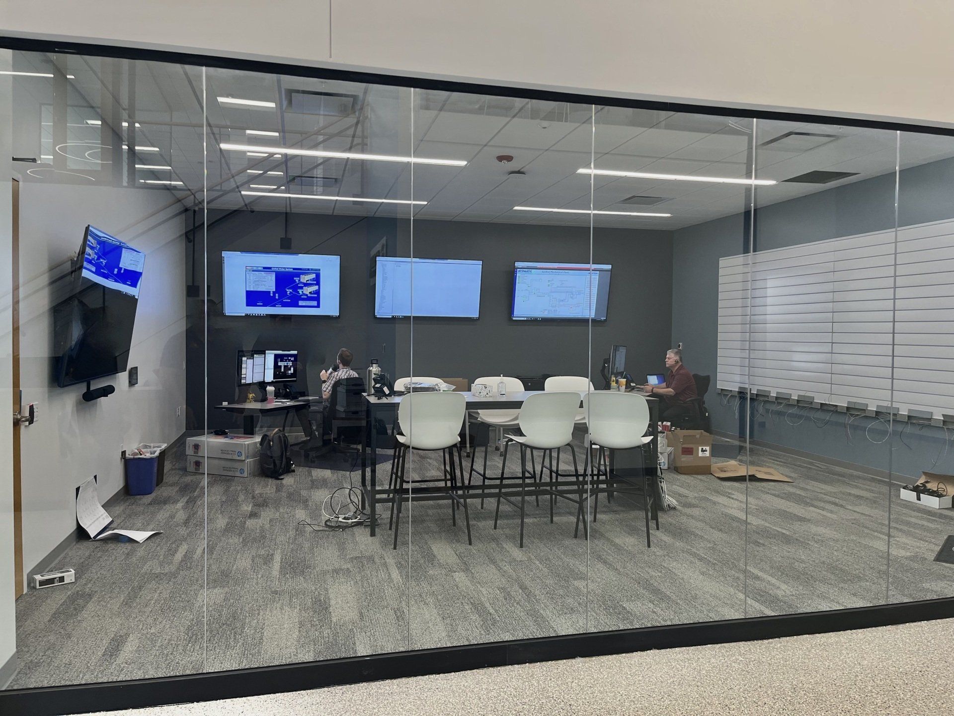 A modern office meeting room with glass walls.  Inside, there is a table with chairs, multiple monitors, and people working.