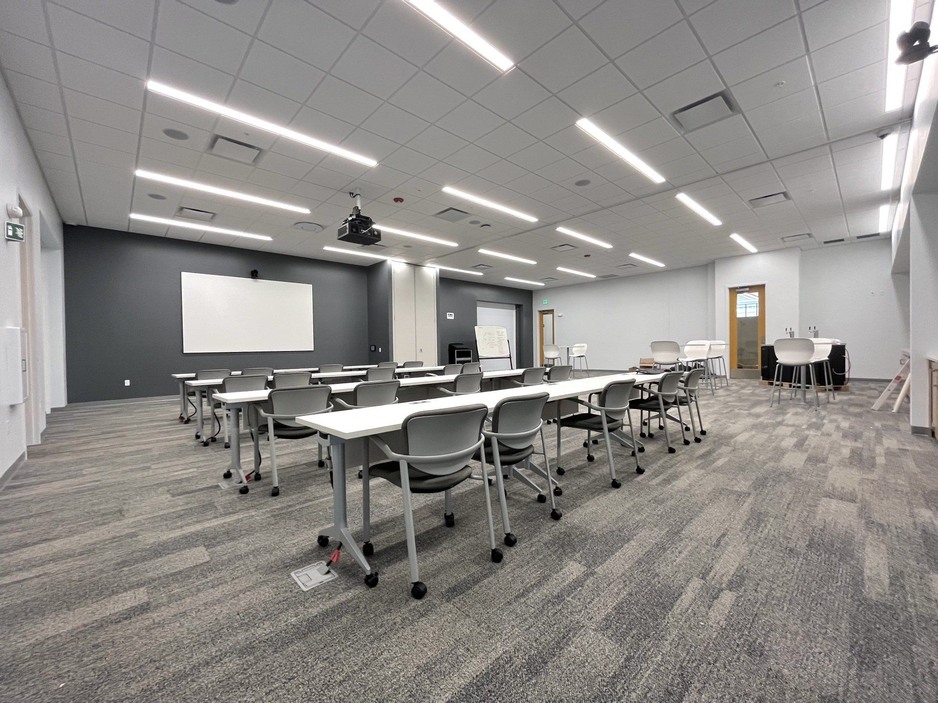 Empty classroom with tables, chairs, whiteboard, and projector. Gray carpet and walls; bright overhead lights.