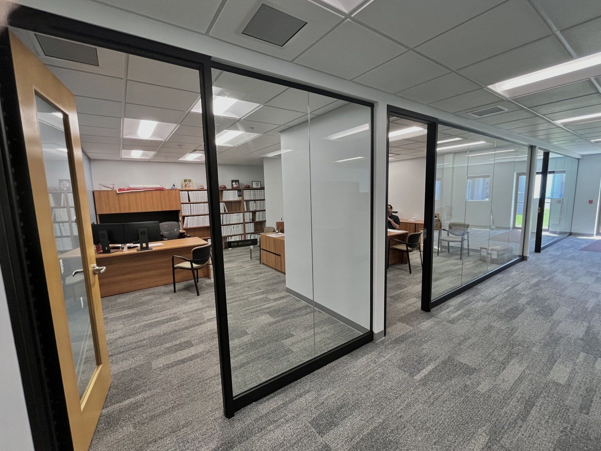 Office space with glass-walled rooms and a door. Desks, computers, and furniture are visible inside.