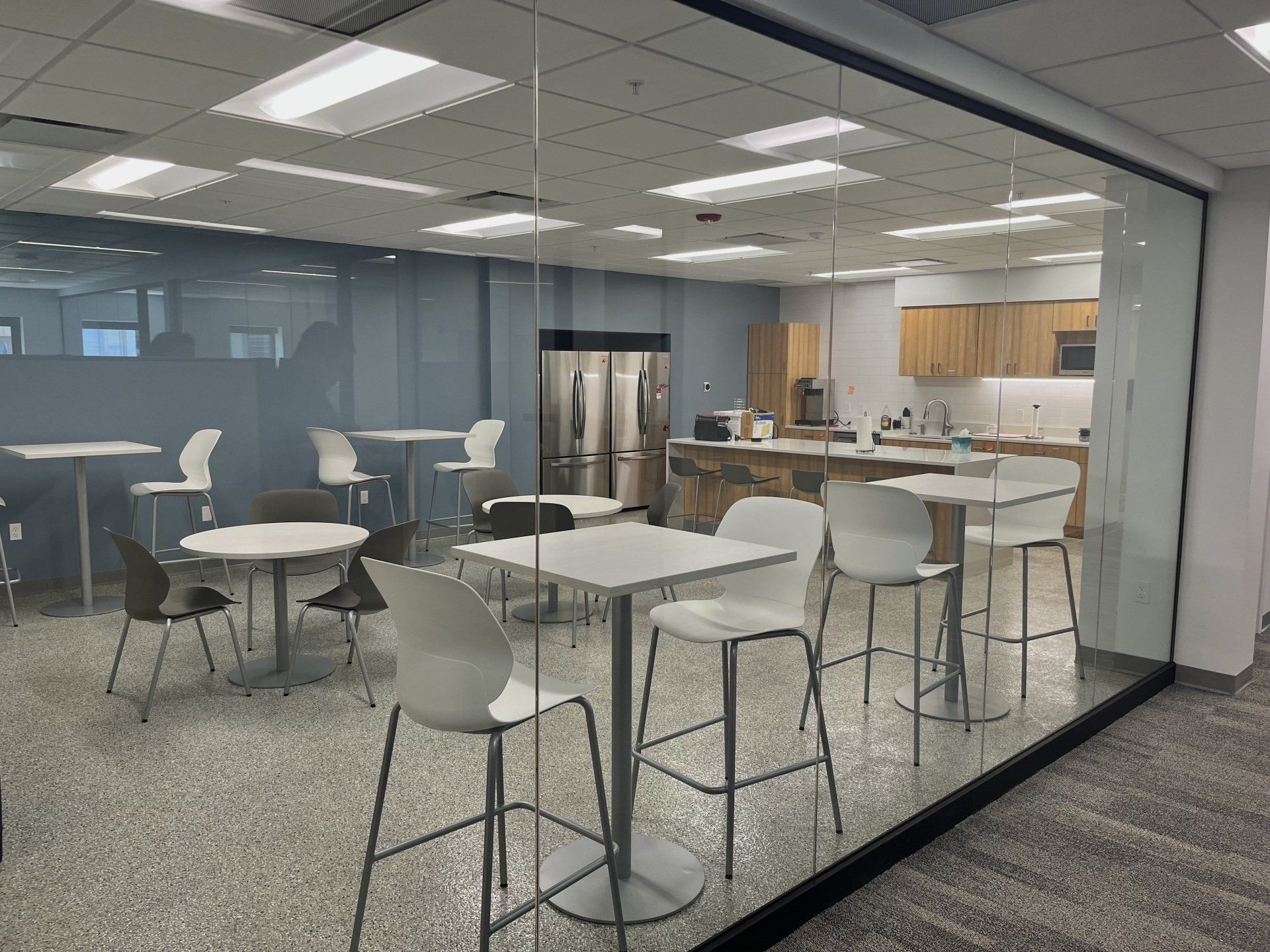 Modern office break room with tables, chairs, and a kitchen area visible through a glass wall.
