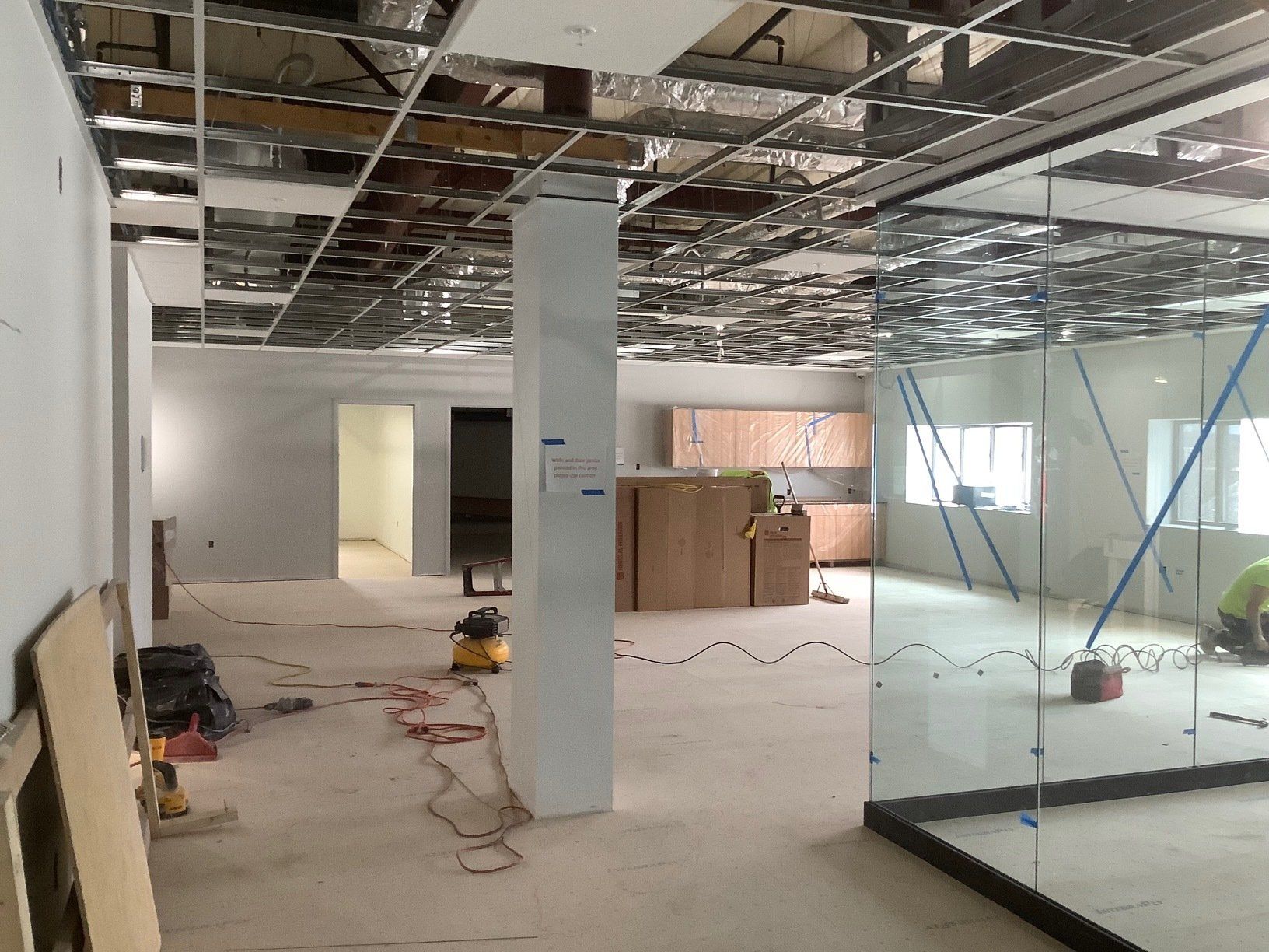 Interior construction site, unfinished ceiling, exposed ductwork. White columns, kitchen cabinets, and glass panels.