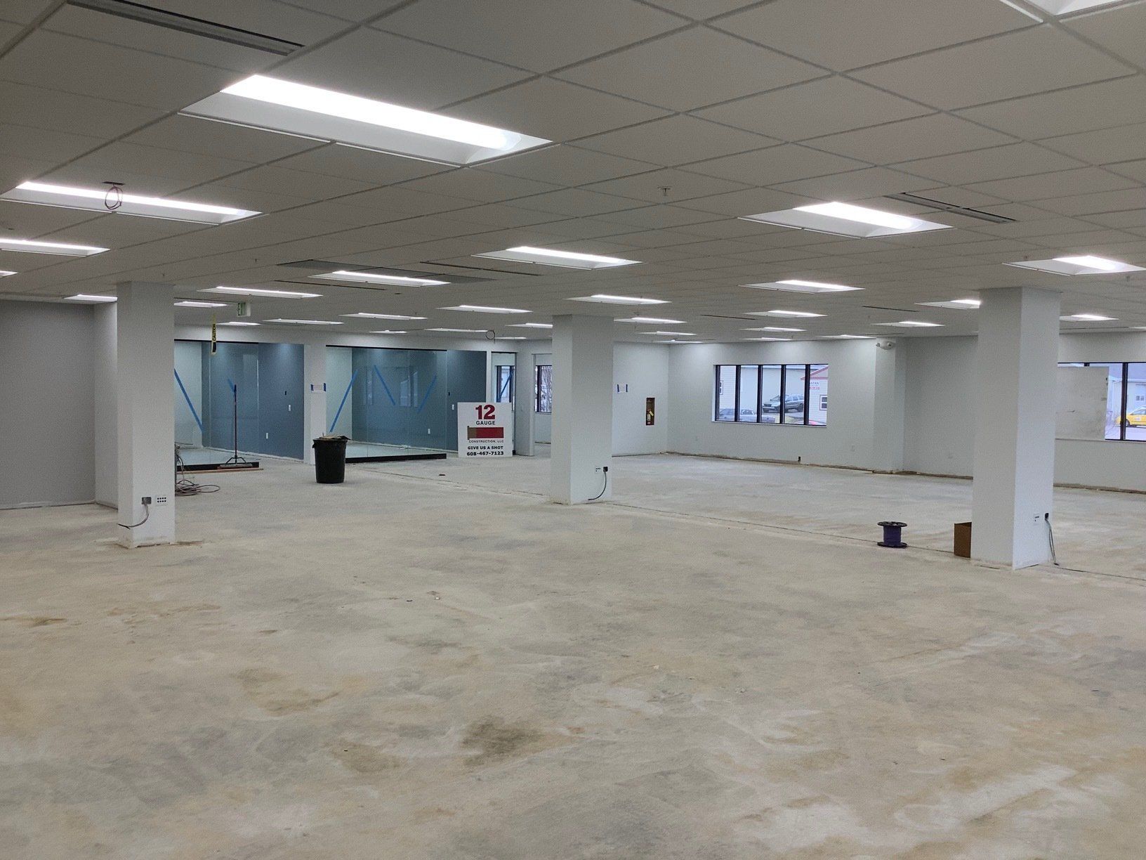 Empty, unfinished commercial interior with white columns, bare concrete floor, and overhead fluorescent lights.