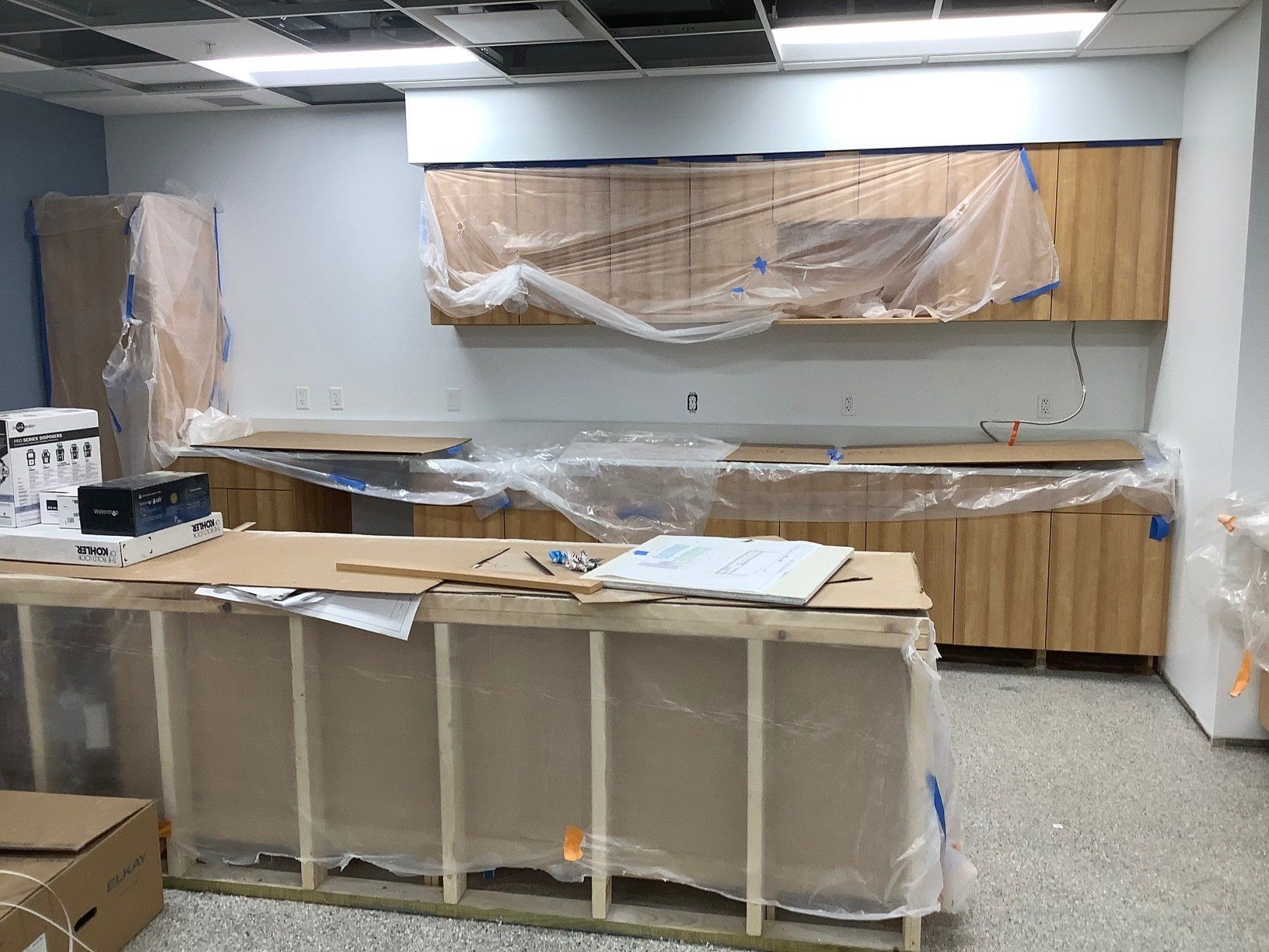 Unfinished kitchen area with wood cabinets, countertop, and a cardboard-covered island, all protected by plastic sheeting.