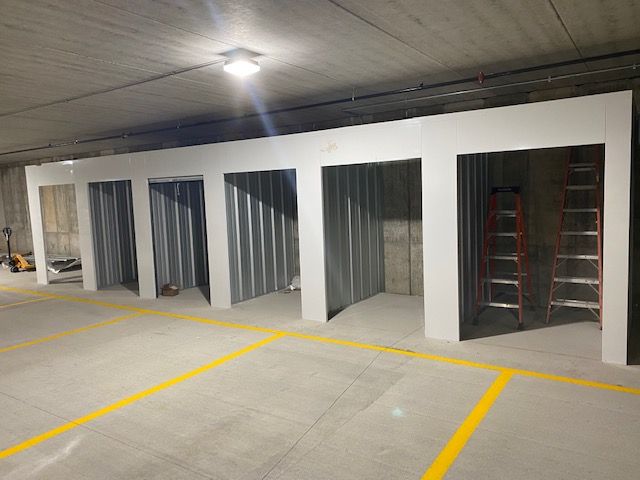 Storage units in a garage with white door frames, grey walls, and yellow floor lines. A red ladder leans inside a unit.