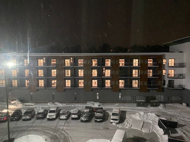 Snowy nighttime view of a multi-story building with lit windows and a parking lot with cars covered in snow.