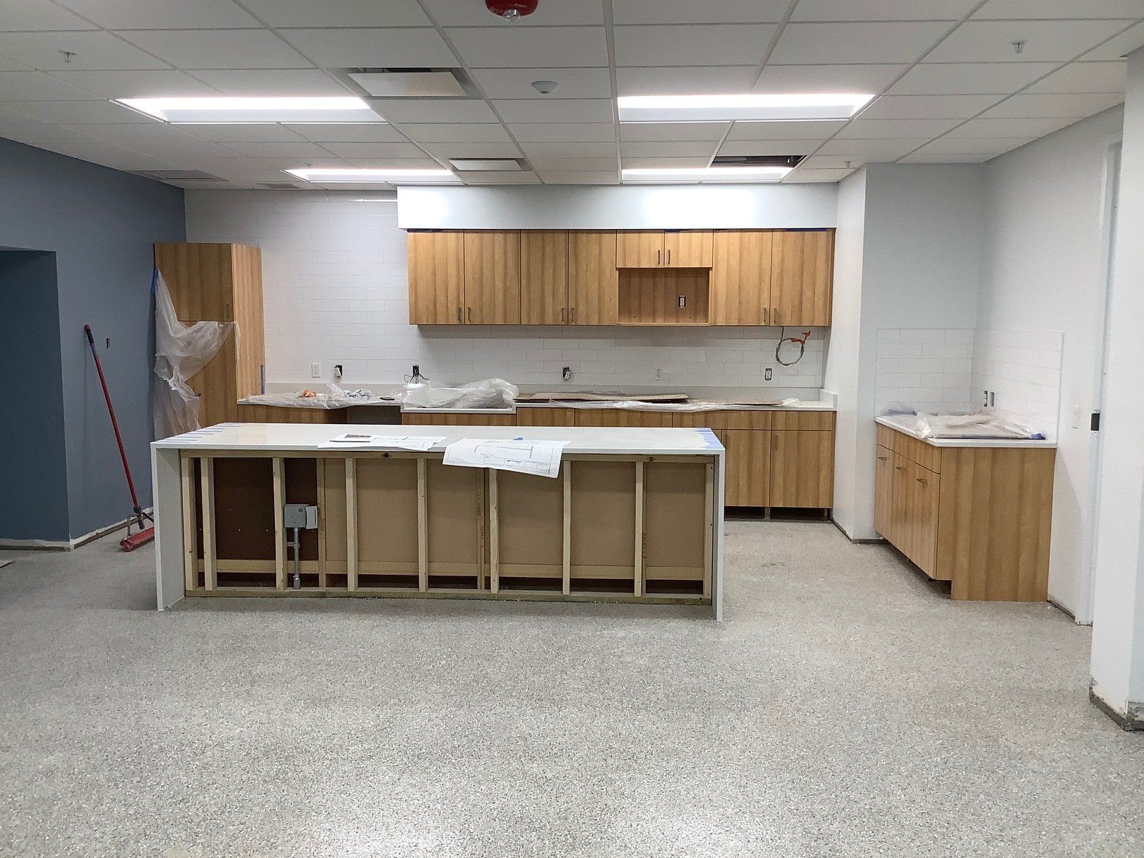 Interior kitchen under construction, with wooden cabinets, island, and speckled flooring. Blue and white walls, unfinished.
