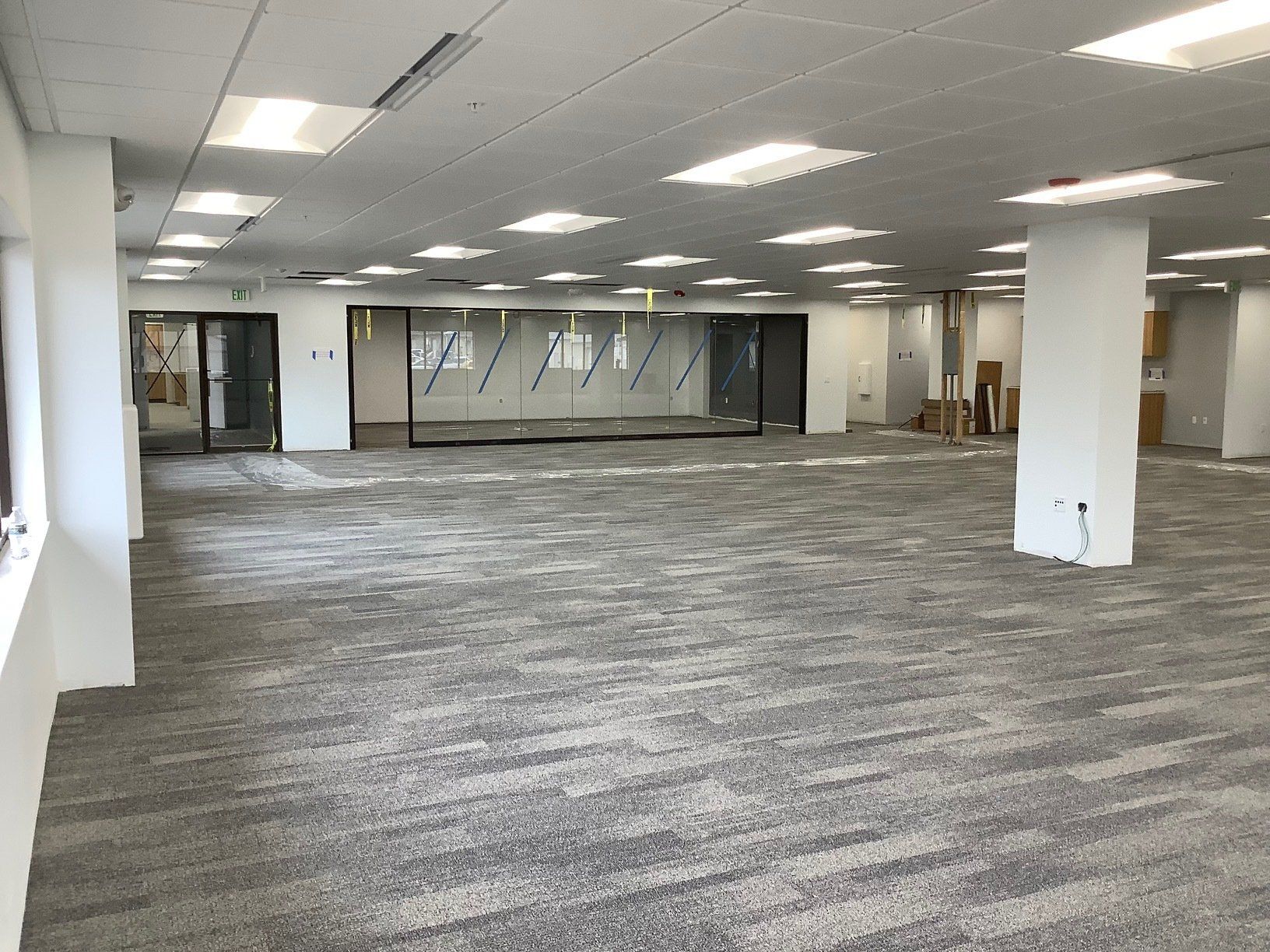 Empty office space with gray carpet, white walls, and glass doors. Fluorescent lights illuminate the room.