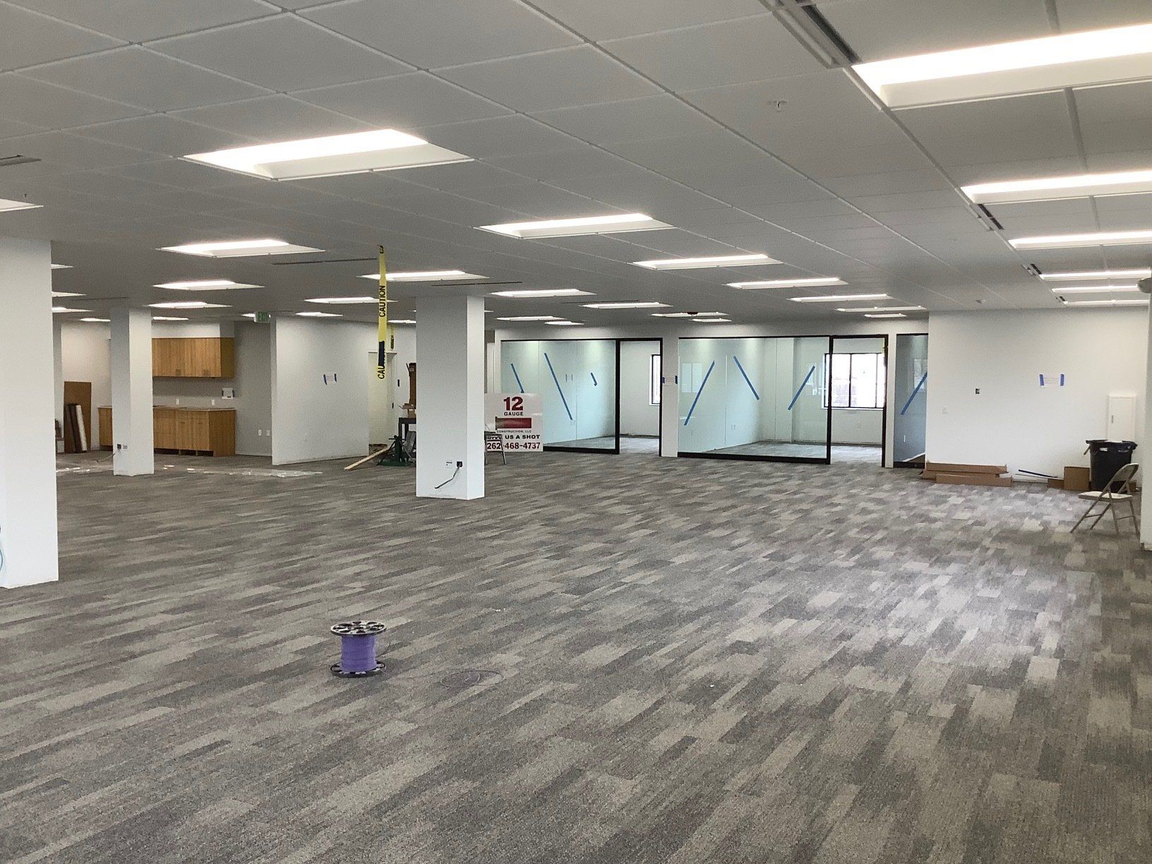 Empty office space with gray carpet, white walls, and glass partitions. Fluorescent lights illuminate the room.