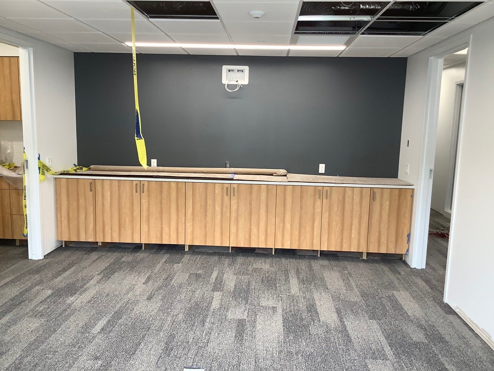 Office space under construction: gray accent wall, wooden cabinets, carpeted floor, exposed ceiling tiles.