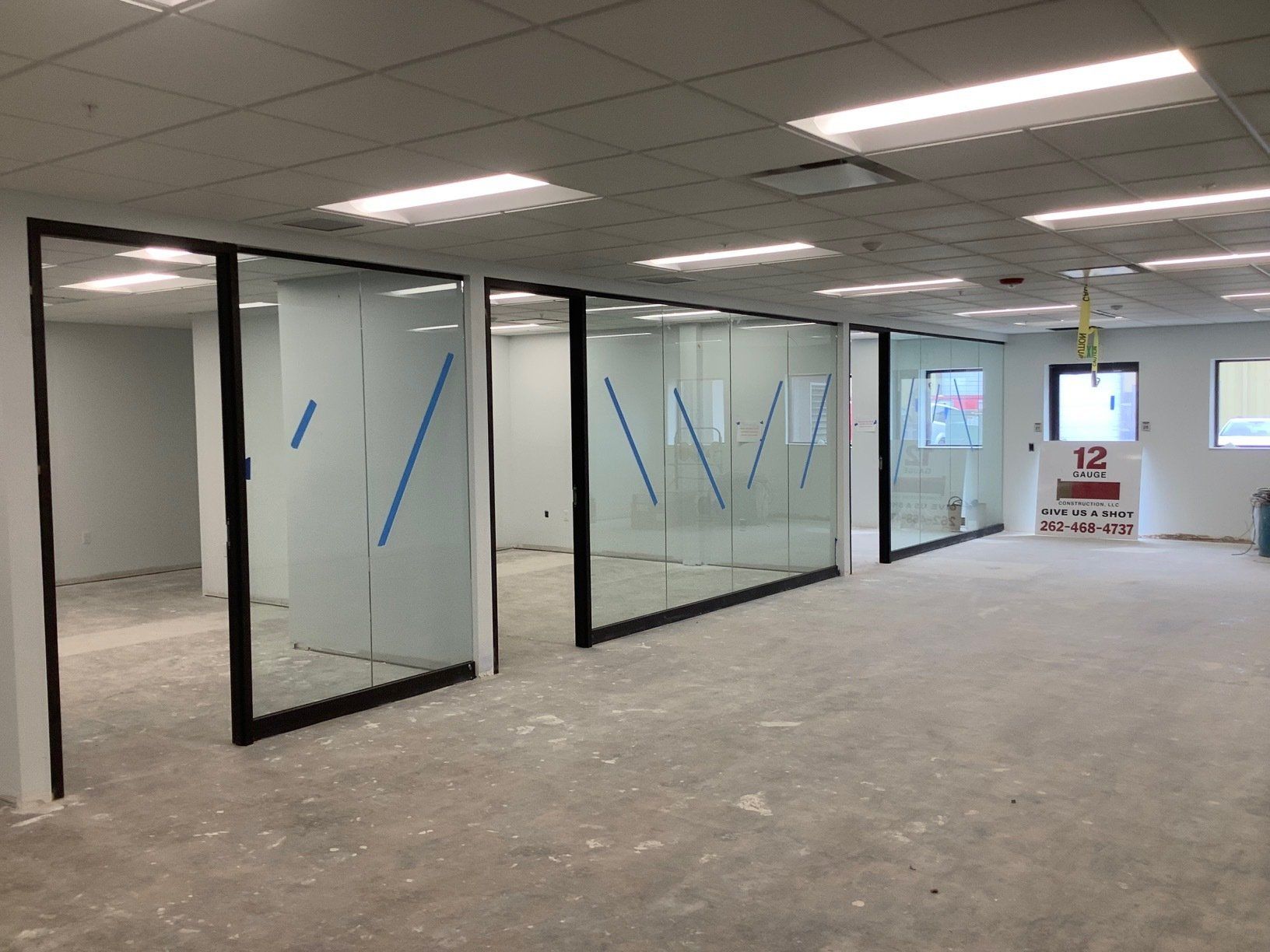 Empty office space with glass-walled rooms, black frames, and overhead lighting. The floor is covered in debris.