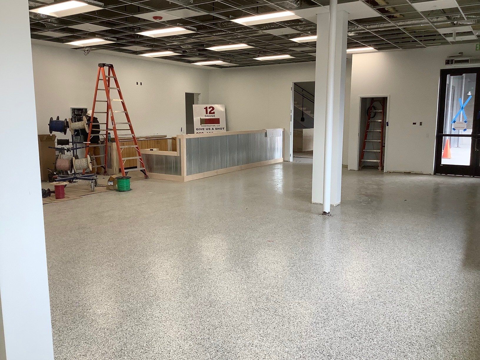 Interior of a construction site with concrete flooring, walls, and a partially finished ceiling. Ladders and tools are present.