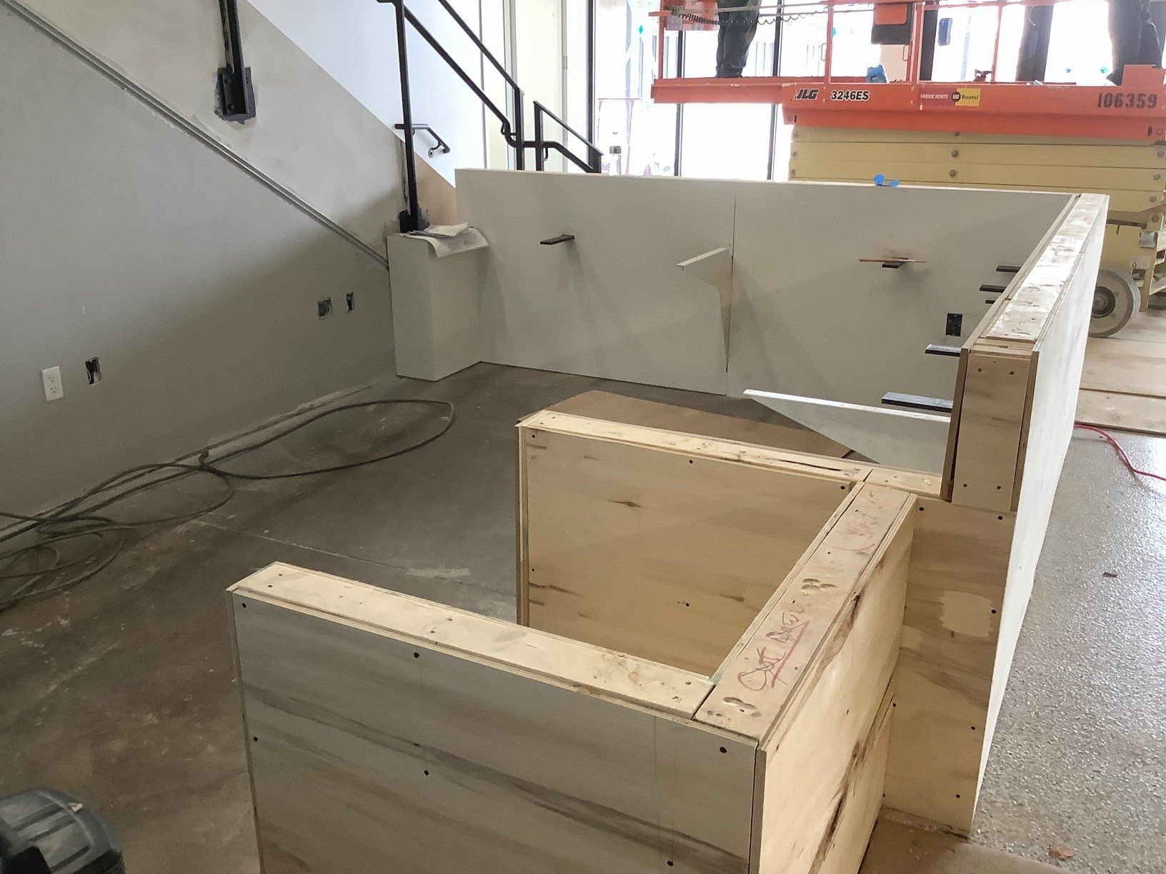 Wooden structure under construction, possibly a counter or barrier. Interior setting with concrete floor, drywall, and lift in background.