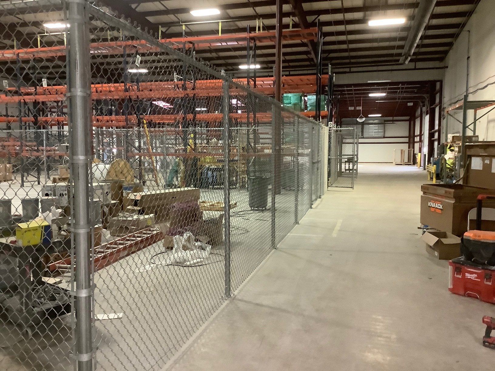 Warehouse interior with a chain link fence enclosing an area with equipment and inventory. A long hallway extends to the right.