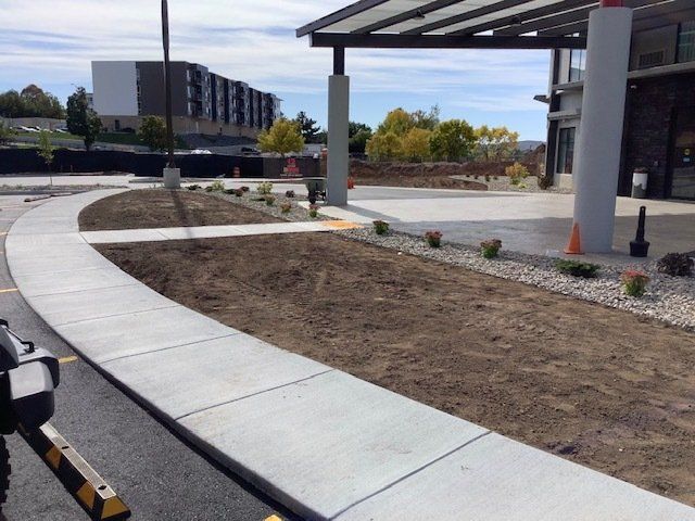 Concrete sidewalk curves in front of a building with a canopy, empty flower beds, and a building in the background.