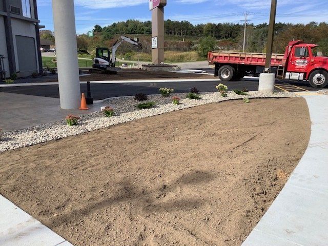 Landscaped area with dirt, gravel, plants, and a truck near a building with a large pillar.