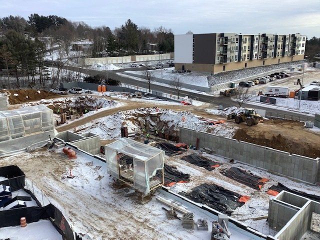 Construction site covered in snow, with completed building in the background. Heavy machinery and foundation work visible.