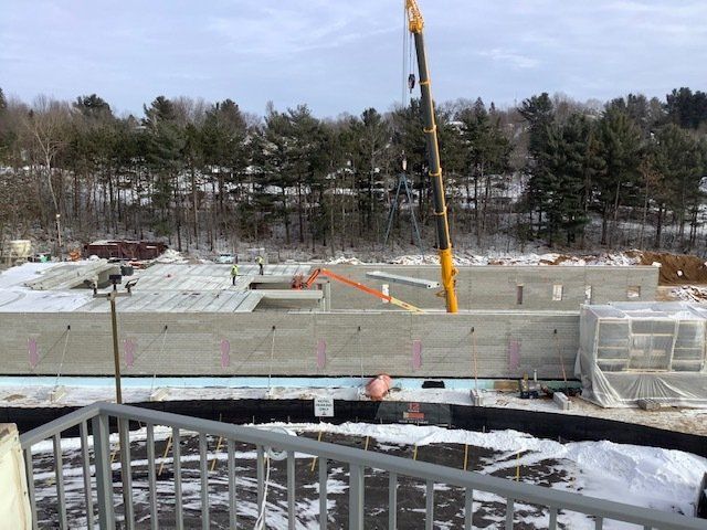 Construction site with a crane lifting a concrete wall. Snow covers the ground, trees in the background.