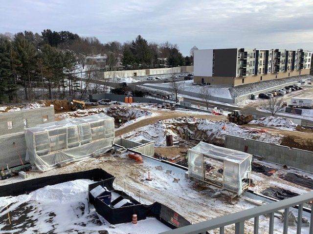 Construction site covered in snow, with buildings, fences, and machinery. Overcast sky.