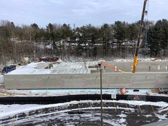 Construction site with crane lifting a concrete wall section. Snowy ground, trees in the background.