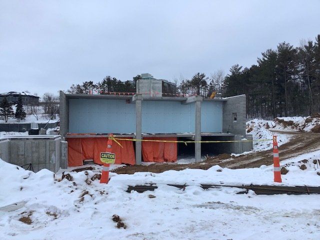 Construction site with snow. Concrete structure with orange barriers, caution tape, and a dirt road. Trees in background.