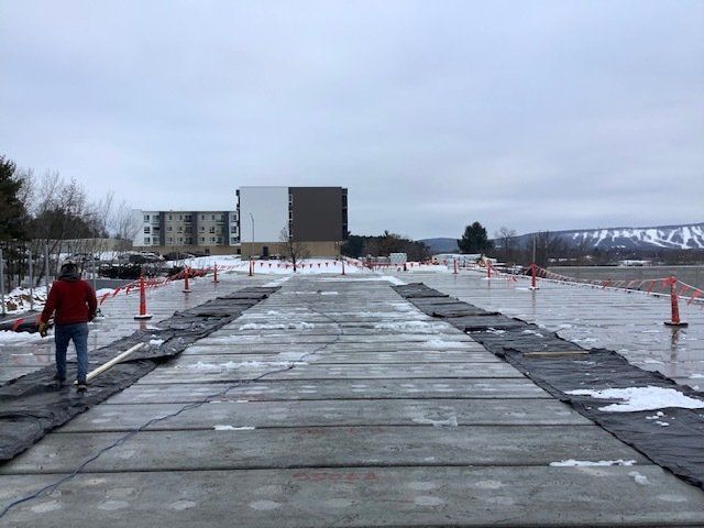 Man walks on concrete slabs laid across a construction site. Red cones and a building are visible. Overcast day.