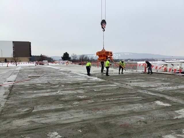 Construction workers guiding machinery suspended by a crane on a concrete slab in a snowy outdoor setting.