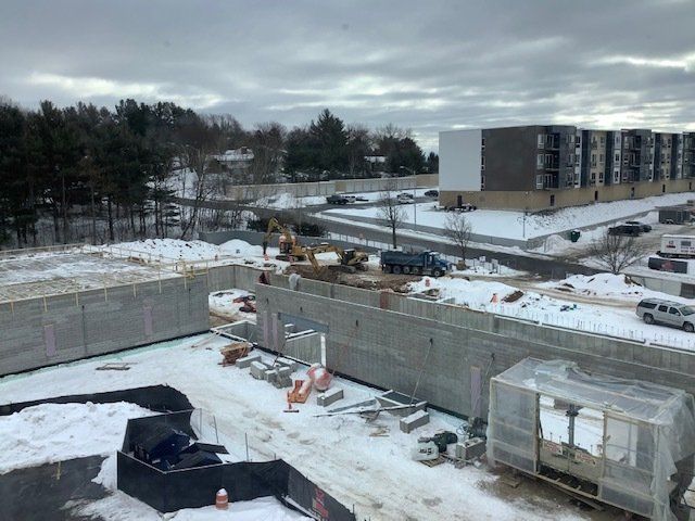 Construction site with snow, concrete walls, and a multi-story building under cloudy skies.