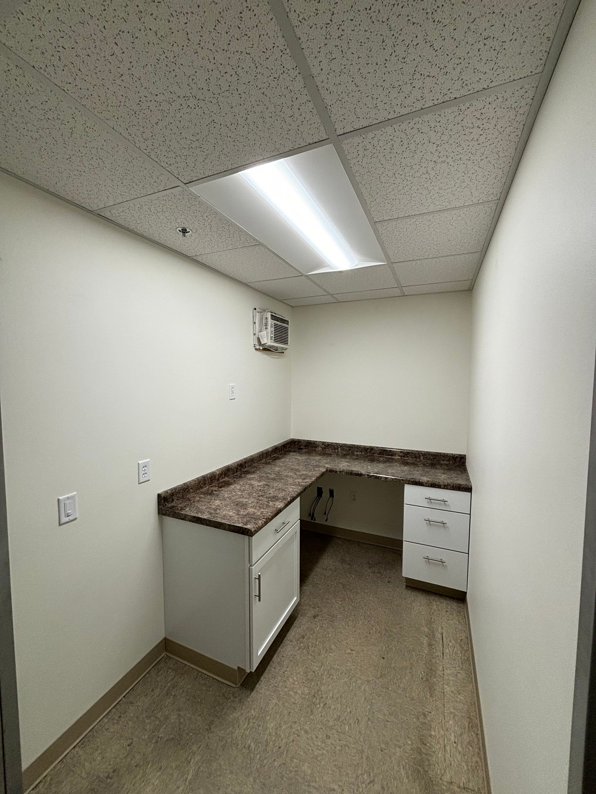An empty office with a built-in desk, cabinets, and a rectangular light fixture. Beige walls, speckled flooring, and a textured ceiling.