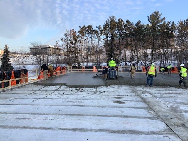 Construction workers pouring and leveling concrete on a bridge deck, snowy surroundings.