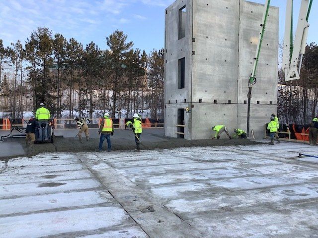 Construction workers pouring concrete at a building site with a concrete structure. Snowy ground, trees in the background.